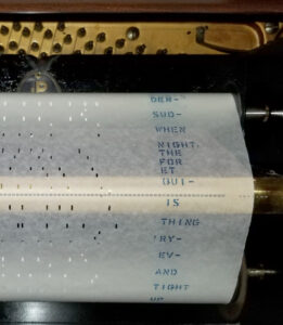 Inside a piano showing a roll of white paper with tiny holes. Along the edge of the paper are the printed lyrics of the song.
